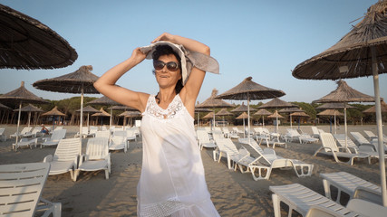 Fototapeta na wymiar Sexy woman in white dress and hat walking on empty beautiful beach with straw umbrellas and beds