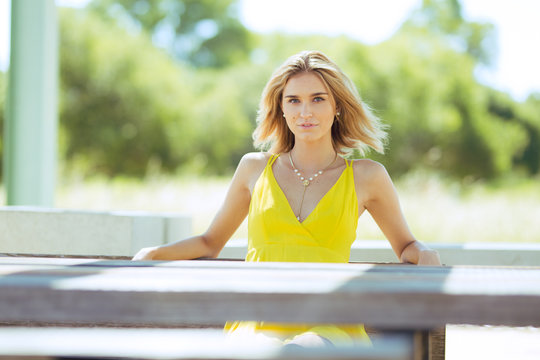 Attractive young blonde Caucasian woman in thin yellow sun dress posing outside on wooden table