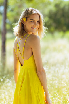 Attractive young blonde Caucasian woman in thin yellow sun dress with flowers in hair posing outside near grass and trees