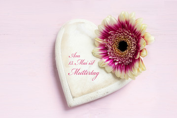 Fototapeta na wymiar white painted wooden heart shape and a flower head on a bright pink background, german text Alles Liebe zum Muttertag, top view from above with copy space