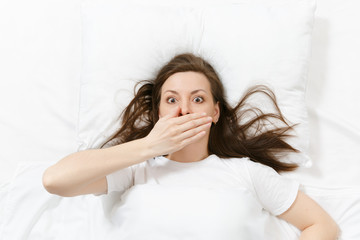 Top view of head of tired brunette young woman lying in bed with white sheet, pillow, blanket. Shocked female cover mouth with hand, spending time in room. Rest, relax, good mood concept. Copy space.