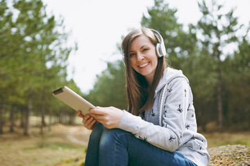 Young smiling beautiful woman in casual clothes with headphones sitting on stone using tablet pc computer in city park or forest on green blurred background. Student lifestyle, leisure concept.