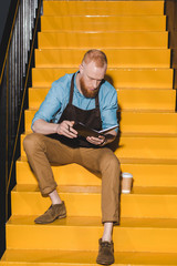 young male barista in apron holding textbook and sitting on stairs with disposable cup of coffee