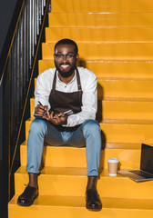 young smiling african american male barista in apron holding textbook and sitting on stairs with laptop and paper cup of coffee