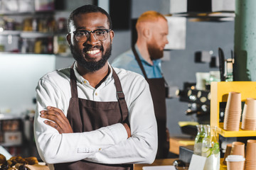 portrait of young smiling african american male barista in apron with colleague standing behind
