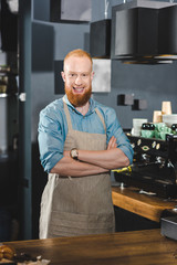 handsome young bearded barista standing with crossed arms and smiling at camera in coffee shop