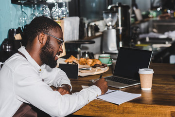smiling young african american barista in eyeglasses taking notes and using laptop with blank screen in cafe