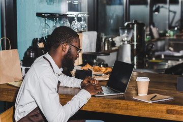 smiling young african american barista in eyeglasses using laptop in coffee shop