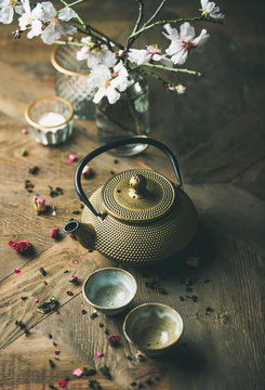 Traditional Asian tea ceremony arrangement. Golden iron teapot, cups, dried rose, candles and almond blossom flowers over vintage wooden table background, selective focus