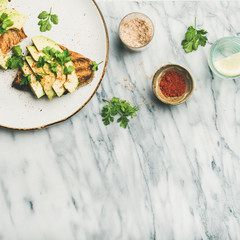 Healthy vegan breakfast or lunch. Flat-lay of avocado toast with seasoning and glass of lemon water over marble background, top view, copy space, square crop. Clean eating, detox, dieting food concept