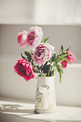 A bouquet of multicolored peonies in a vase in the form of watering can is in a white Sunny living room.