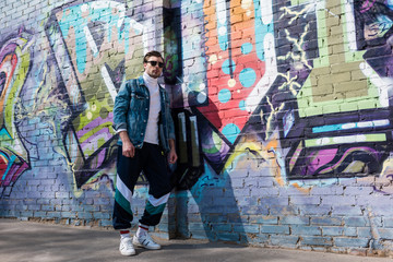 stylish young man in vintage denim jacket and track pants leaning on brick wall with graffiti