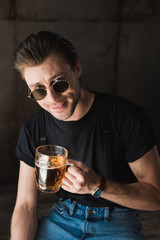 happy young man in black t-shirt and sunglasses holding mug of beer and looking at camera