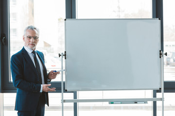 handsome senior businessman in eyeglasses pointing at blank whiteboard and looking at camera