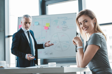 Obraz premium beautiful businesswoman smiling at camera while senior businessman standing at whiteboard behind