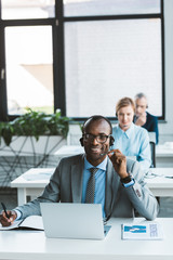 young african american businessman in eyeglasses and headset smiling at camera while working with laptop in office