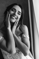 Happy adorable bride smiles tender posing in a wedding dress before a bright window