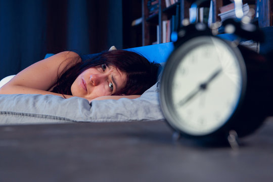 Picture of brunette with insomnia lying on bed next to alarm clock at night