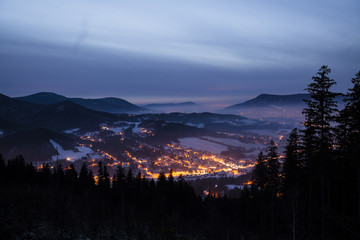 Night beautiful scene of winter landscape of shining village surrounded by mountains and deep spruce tree forest in fog during twilight. Creative long exposure photography with headlight motion blur.