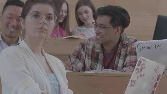 Positive studens laughing during studing in the university classroom, sitting on wooden desks. Working with laptops, wearing casual clothes and eyeglasses. Smiling, feeling happy.