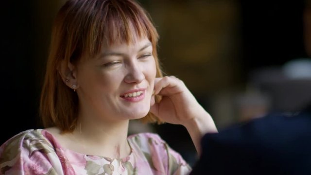 Tilt up of cheerful young woman in floral dress laughing and drinking tea while on date with unrecognizable man in cafe