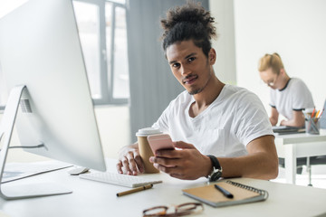young african american man looking at camera while using smartphone and desktop computer in office