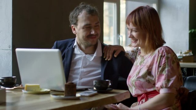 Medium shot of happy young woman in floral dress and cheerful man in jacket sitting at tablet in cafe and joking while talking to someone on video call via laptop