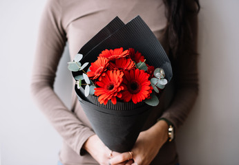 Very nice young woman holding a fresh blossoming flower bouquet of vivid crimson red gerbera mini and eucalyptus on the grey wall background