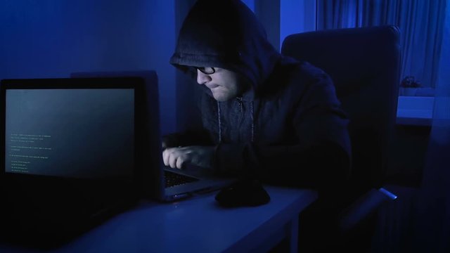 Funny 4k footage of male pretends to be hacker. Geeky man in hood working on laptop at night