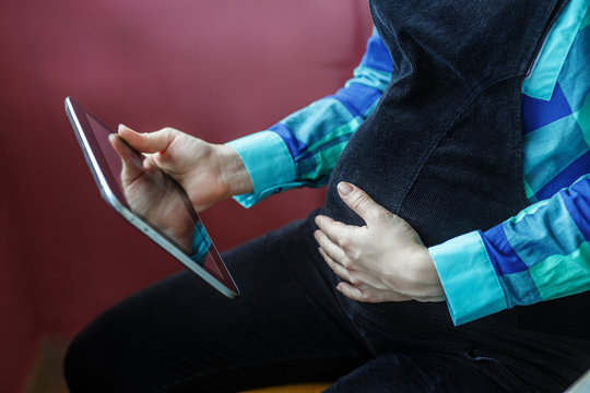 A pregnant woman is sitting with a tablet in her hands. The concept of a lifestyle, motherhood, work, shopping, the Internet.