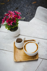Cup of cappuccino coffee with syrup and pink flower on white table in the garden.
