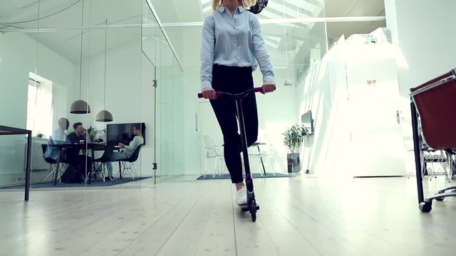 Young businesswoman laughing while riding a scooter during her break around a large modern office
