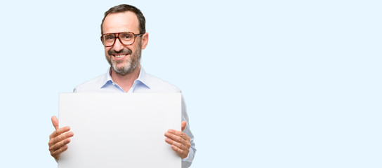 Middle age man with glasses holding blank advertising banner, good poster for ad, offer or announcement, big paper billboard isolated over blue background