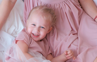 Little charming blond girl plays hide and seek with her mom in pink dress laying on bed.