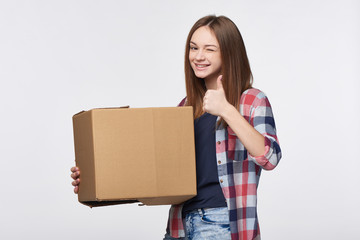 Delivery, relocation and unpacking. Happy playful young woman holding cardboard box winking at camera and gesturing thumb up