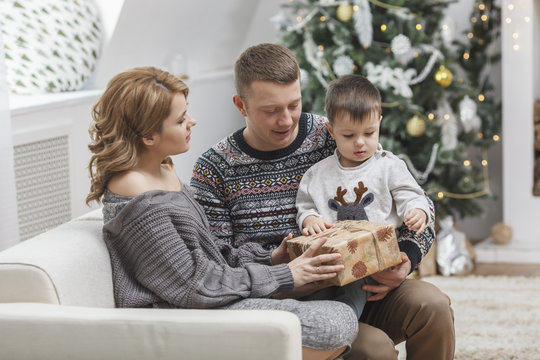 Man looking at woman giving gift to son sitting on sofa at home during Christmas