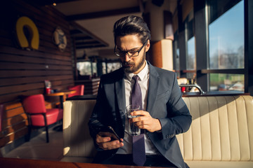 Successful stylish focused handsome bearded businessman in the suit looking at a mobile while holding a glass of water in a cafe or restaurant.