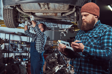 Two bearded brutal mechanics repair a car on a lift in the garage. 