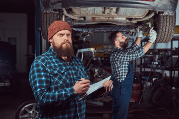 Two bearded brutal mechanics repair a car on a lift in the garage. 