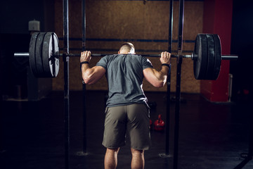 Fototapeta na wymiar Rear view of young short hair focused strong muscular bodybuilder man standing with a heavy barbell behind the neck while preparing for squat exercise.