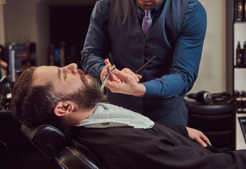 Fototapeta premium Professional hairdresser modeling beard with scissors and comb at the barbershop.
