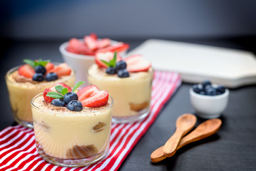 homemade, exquisite dessert tiramisu in glasses decorated with strawberry, blueberry, mint on black wooden table