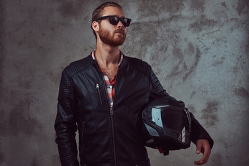 Obraz na płótnie Canvas Portrait of a handsome stylish redhead biker in a black leather jacket and sunglasses, holds motorcycle helmet, posing in a studio.
