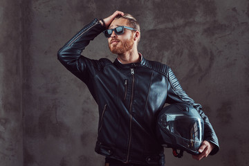 Fototapeta na wymiar Portrait of a handsome stylish redhead biker in a black leather jacket and sunglasses, holds motorcycle helmet, posing in a studio.