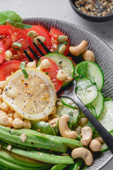 Close up of a healthy vegan lunch bowl with raw avocado, chickpeas, cashew, tomatoes, cucumber, greens and seeds over white kitchen table.