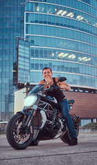Fototapeta na wymiar A handsome brutal biker dressed in a black t-shirt and jeans, sitting on a sports motorcycle, is nostalgic against a skyscraper in a city downtown.