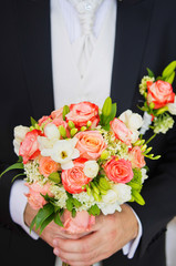 Big bridal bouquet of white and orange fresh flowers in hands of groom in black suit. Vertical color photography.