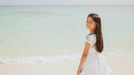 Fototapeta na wymiar A charming philippine schoolgirl in a white dress is walking along a white sandy beach. Enjoying the tropical scenery. Childhood.