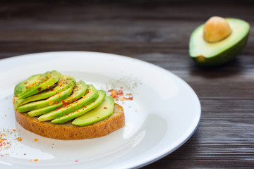 Toast with cutted avocado and spices in white plate on brown wooden background