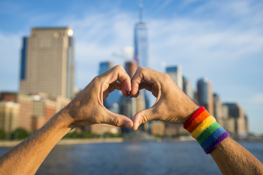 Hands wearing gay pride rainbow sweat band making heart symbol in front of city skyline